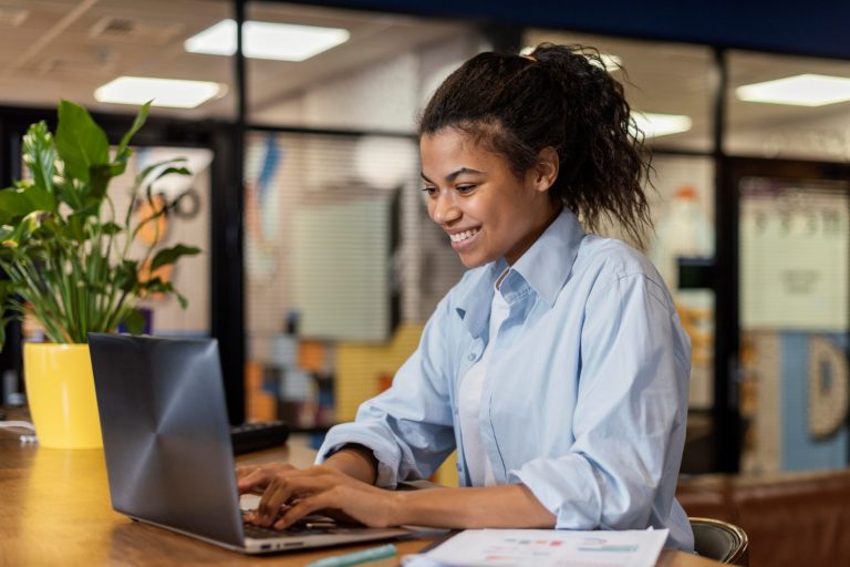 Smiling young woman sitting in front of laptop illustrates blog "SBA 7(a) Loans for Franchises: What You Need To Know"