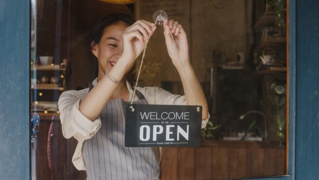 Photo of girl hanging a sign that reads "open" illustrates blog: "Can SBA Loans Be Used To Start a Business?"