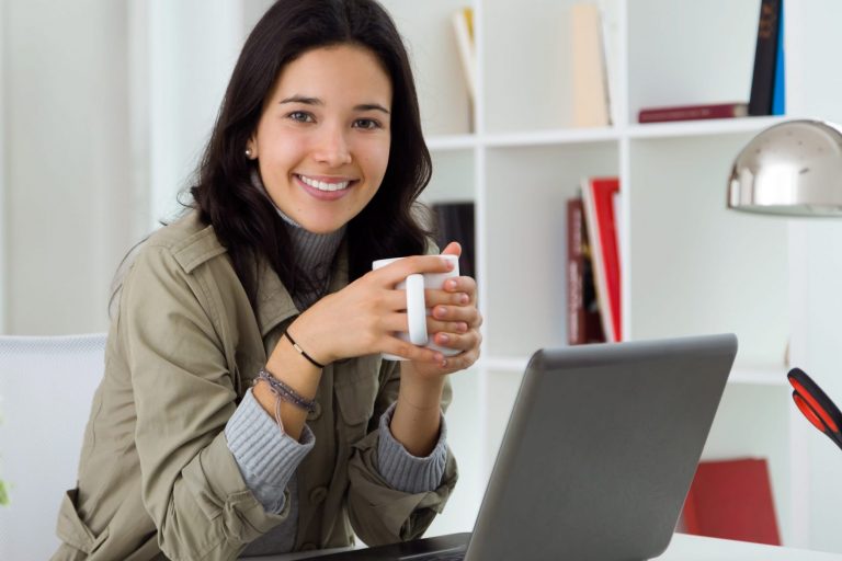 Photo of smiling woman holding coffee mug in front of computer illustrates blog: "Bridge Loans: the Pros and Cons"