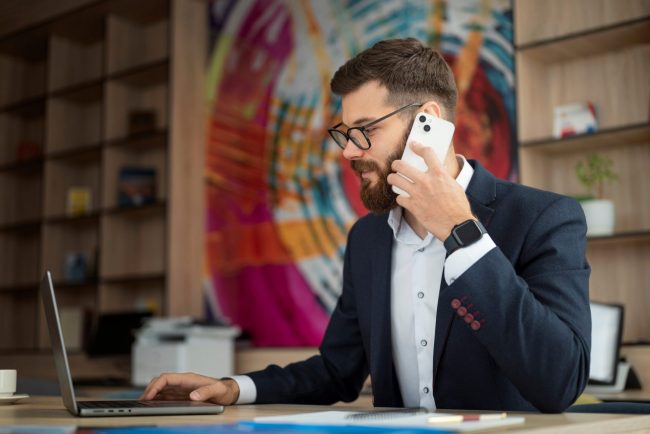 Man sitting at desk working on laptop while talking on cell phone illustrates blog "Are SBA Loans Hard To Get?"