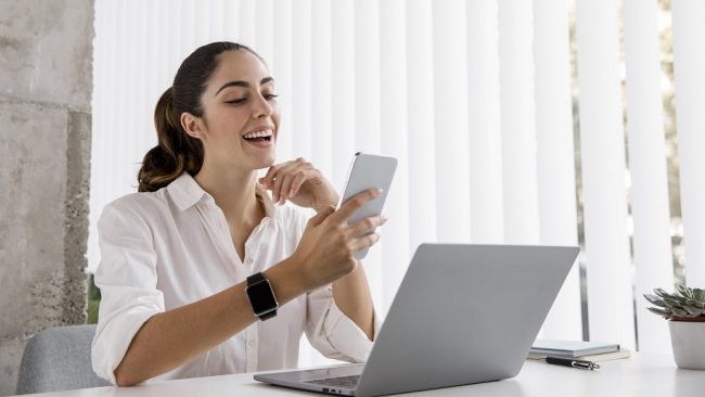 woman-office-freepik Woman in office in front of laptop looking at smartphone illustrates blog "Why Revenue-Based Financing Could Be a Game-Changer for Your SME"