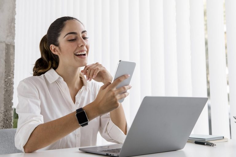 woman-office-freepik Woman in office in front of laptop looking at smartphone illustrates blog "Why Revenue-Based Financing Could Be a Game-Changer for Your SME"